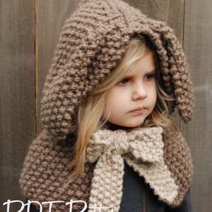 May include: A brown knitted hooded scarf with a matching beige scarf tied in a bow. The scarf is worn by a young girl with blonde hair.