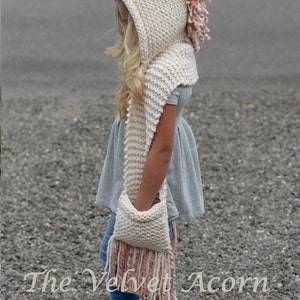 May include: A white knitted unicorn hooded scarf with pink and white yarn accents. The scarf has a pocket and long fringe. The text "The Velvet Acorn Designs" is visible at the bottom of the image.