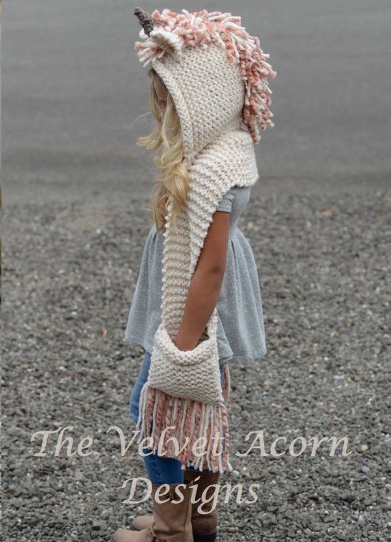 May include: A white knitted unicorn hooded scarf with pink and white yarn accents. The scarf has a pocket and long fringe. The text "The Velvet Acorn Designs" is visible at the bottom of the image.