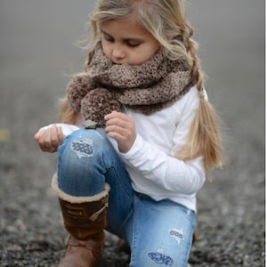 May include: A young girl wearing a brown knit infinity scarf with pom poms, a white long-sleeved shirt, and blue jeans with patches. She is kneeling on a bed of gravel.