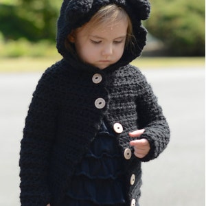 May include: A black crocheted hooded cardigan with bear ears, featuring wooden buttons. The cardigan is worn by a child.