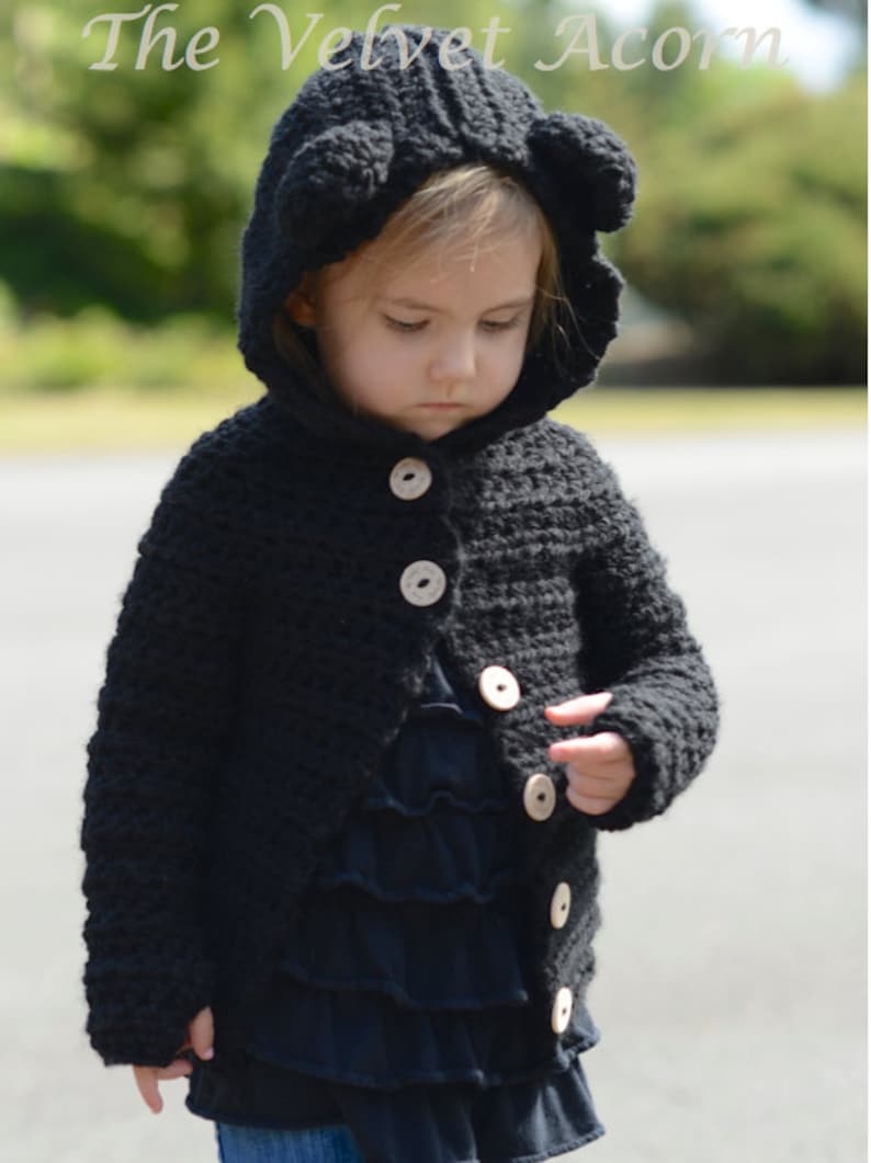 May include: A black crocheted hooded cardigan with bear ears, featuring wooden buttons. The cardigan is worn by a child.