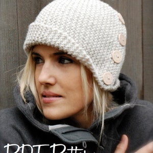 Puede incluir: Un gorro de punto blanco con estilo cloche y cuatro botones de madera en el lateral. El gorro lo lleva una persona con el pelo rubio.