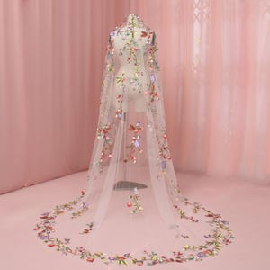 Pode incluir: Um véu de noiva branco transparente com bordados florais coloridos. O véu é drapeado sobre um manequim, mostrando o intrincado design de flores e folhas vermelhas, roxas, verdes e amarelas. A borda do véu também é adornada com o mesmo padrão floral.