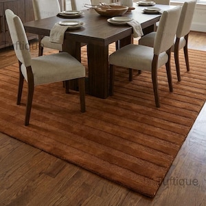 Op de afbeelding: Een eethoek met een donkerhouten tafel en zes gestoffeerde stoelen. De tafel is gedekt met borden en servetten. Een roestkleurig tapijt bedekt de vloer en de kamer wordt verlicht door een kroonluchter.