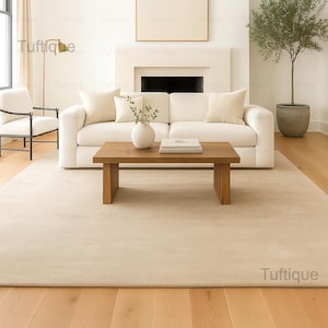 Può includere: Un tappeto rettangolare beige chiaro in un soggiorno. Il tappeto è posto sotto un divano bianco, un tavolino in legno e una sedia bianca. La stanza ha pavimenti in legno e un design minimalista. Il tappeto è di colore neutro.