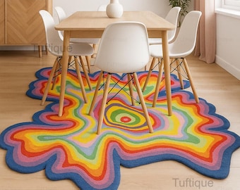 Puede incluir: Una colorida alfombra abstracta con un diseño único y fluido. La alfombra presenta capas concéntricas de colores vibrantes, incluyendo azul, verde, amarillo, naranja y rojo, con un borde azul. La alfombra está colocada debajo de una mesa de madera y sillas blancas.