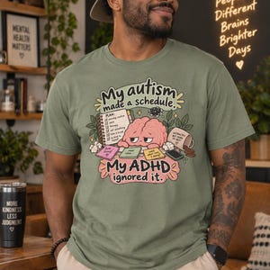 May include: An olive green t-shirt featuring a graphic illustration of a pink brain with a tired expression. The text reads: My autism made a schedule. My ADHD ignored it. The design includes a checklist, sticky notes, and a spilled coffee mug.