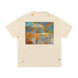 Può includere: T-shirt color crema con una colorata stampa d'arte astratta sul retro. L'opera raffigura una struttura a forma di casa con un sole rosso e forme geometriche. La maglietta presenta schizzi di vernice in vari colori.