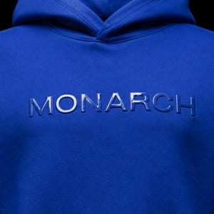 May include: A royal blue hoodie with the word "MONARCH" embossed across the chest. The hoodie features a hood and a soft, comfortable material. The text is in a lighter shade of blue.