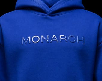 May include: A royal blue hoodie with the word "MONARCH" embossed across the chest. The hoodie features a hood and a soft, comfortable material. The text is in a lighter shade of blue.