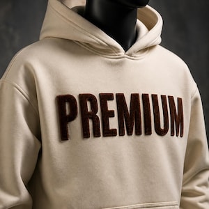 May include: Cream-colored hoodie with a kangaroo pocket and a hood. The word "PREMIUM" is embroidered in a dark brown, textured font across the chest. The hoodie is displayed on a mannequin.
