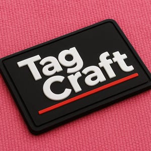 May include: A black rectangular patch with the white text "Tag Craft" and a red horizontal line. The patch is set against a pink textured background.