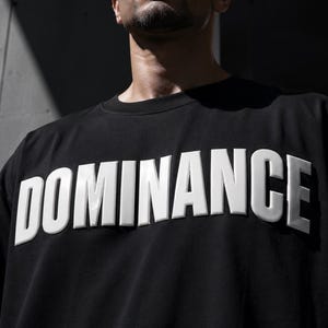 May include: Black crew neck t-shirt with the word "DOMINANCE" in large, raised white letters across the chest. The shirt is a solid black color and appears to be made of a soft material. The image is a close-up shot.