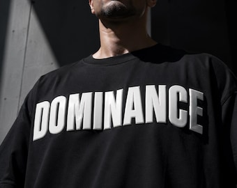 May include: Black crew neck t-shirt with the word "DOMINANCE" in large, raised white letters across the chest. The shirt is a solid black color and appears to be made of a soft material. The image is a close-up shot.