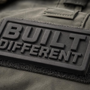 May include: Close-up of a dark gray rectangular patch with the words "BUILT DIFFERENT" in bold, raised letters. The patch is attached to a green fabric background. The patch has a textured surface.