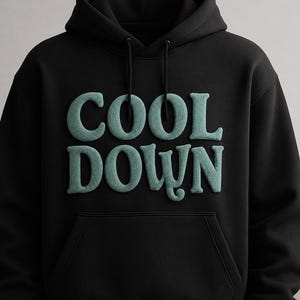 Puede incluir: Sudadera con capucha negra con la frase "COOL DOWN" en letras turquesas texturizadas en el pecho. La sudadera tiene un bolsillo delantero y una capucha con cordón. El diseño es simple y moderno.