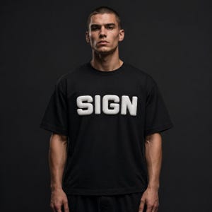 Op de afbeelding: Zwart T-shirt met ronde hals en het witte woord "SIGN" over de borst. De tekst heeft een verhoogde, 3D-uitstraling. Het shirt is gemaakt van een zacht materiaal en is geschikt voor vrijetijdskleding.