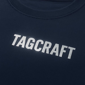 Puede incluir: Sudadera azul marino con la palabra "TAGCRAFT" impresa en plata en la parte delantera. El texto está en negrita, con una fuente sans-serif. La sudadera tiene cuello redondo y parece estar hecha de un material suave.