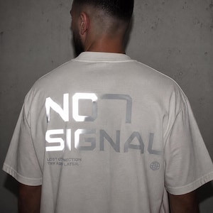 Könnte beinhalten: Cremefarbenes T-Shirt mit den Worten "NO SIGNAL" in reflektierendem silbernen Schriftzug auf dem Rücken. Darunter stehen die Worte "LOST CONNECTION TRY AGAIN LATER" und eine Weltkugel-Grafik. Das Shirt hat kurze Ärmel.