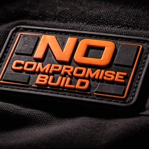 Può includere: Toppa rettangolare nera con le parole "NO COMPROMISE BUILD" in lettere arancioni. La toppa ha un bordo nero ed è posta su uno sfondo nero strutturato.