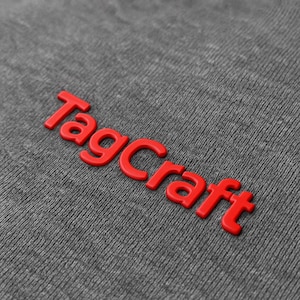 May include: A close-up shot of the word "TagCraft" in bold, red, raised lettering. The letters are 3D and stand out against a textured, dark grey fabric. The lighting creates subtle shadows, enhancing the visual impact of the text.