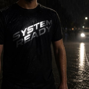 May include: Black t-shirt with the text "SYSTEM READY" in white, worn in the rain. The image shows a person standing on a wet street at night, with car headlights in the background. The shirt appears to be made of a moisture-wicking material.