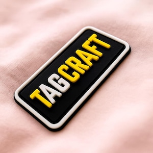 May include: A rectangular patch with the word "TAGCRAFT" in white and yellow raised letters on a black background. The patch has a white border and is set against a pink fabric.