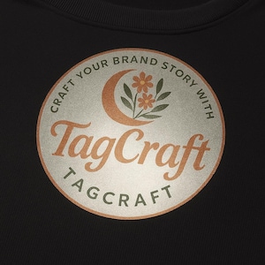 May include: Black sweatshirt with a circular logo featuring the text "TagCraft" in orange script. The logo also includes a crescent moon and flowers, with the words "Craft Your Brand Story With" around the edge.