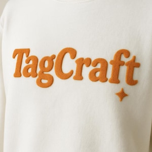May include: Off-white sweatshirt featuring the orange text "TagCraft" in a textured, raised font. A small orange star is positioned below the text. The sweatshirt has a crew neck and a minimalist design.