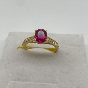 May include: A gold ring featuring a large, oval-cut, magenta gemstone. The band is also gold and is embellished with small, clear gemstones. The ring is set against a white background.