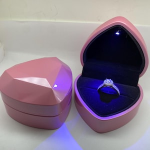 May include: A pink heart-shaped ring box, open to reveal a diamond ring. The box is lit with a blue light, and the ring is set against a black velvet interior. A second, closed box is next to it.