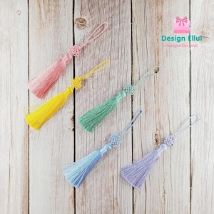 Peut inclure: Cinq pompons colorés, rose, jaune, vert, bleu et violet, chacun avec un nœud décoratif et une boucle. Les pompons sont disposés sur une surface en bois clair. Le texte "Design Ellut" est visible en haut à droite.