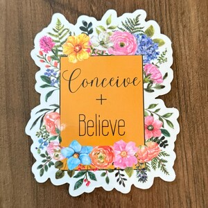 May include: A sticker with a floral border and the words "Conceive + Believe" in black script on an orange background. The flowers include pink, yellow, blue, and orange blooms.