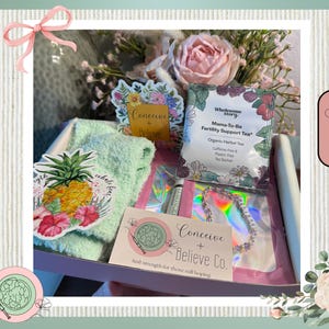 May include: A gift box with a variety of items, including a tea bag, a bracelet, and a sticker with the words "Conceive + Believe". The box also contains a green towel and a sticker with a pineapple design. The box is labeled "The Conceive + Believe Box".