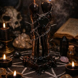 Puede incluir: Dos velas cil&iacute;ndricas negras y altas est&aacute;n colocadas una al lado de la otra, ambas encendidas con llamas brillantes. Una cadena de metal plateado envuelve ambas velas, asegurando un peque&ntilde;o dije negro en forma de coraz&oacute;n en el centro. Varios pinchos met&aacute;licos afilados sobresalen de los lados de los pilares de cera, que se asientan sobre una base cuadrada oscura.