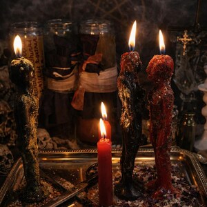 Puede incluir: Tres velas con forma humana en negro y rojo, cada una con una llama encendida, se alzan sobre una bandeja met&aacute;lica junto a una vela de pilar roja m&aacute;s peque&ntilde;a y unas tijeras de metal. Las velas tienen textura de hierbas y purpurina, rodeadas de p&eacute;talos.