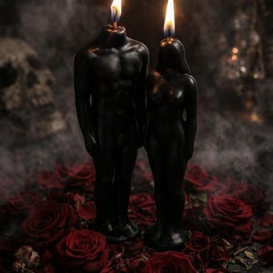 May include: Two black wax candles shaped like human figures, one with a broader torso and one with a slender frame, both standing upright with lit flames at the top, surrounded by scattered deep red rose petals on a dark surface.