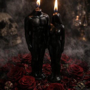 Puede incluir: Dos velas de cera negra con forma de figuras humanas, una con un torso m&aacute;s ancho y otra con una complexi&oacute;n esbelta, ambas de pie con las mechas encendidas. Las velas est&aacute;n rodeadas por numerosas flores de rosa de color rojo intenso.
