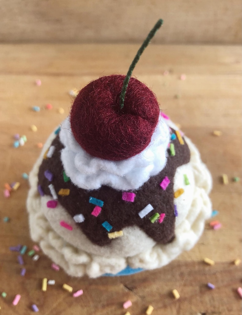 May include: A felt ice cream cone with white frosting, chocolate sauce, and colorful sprinkles topped with a red felt cherry.