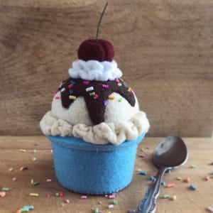 May include: A felt ice cream cone with a cherry on top, chocolate sauce, and sprinkles. The cone is blue and the ice cream is white. The ice cream is sitting on a wooden surface with a silver spoon.