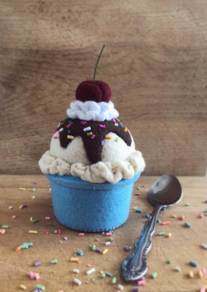 May include: A felt ice cream cone with a cherry on top, chocolate sauce, and sprinkles. The cone is blue and the ice cream is white. The ice cream is sitting on a wooden surface with a silver spoon.