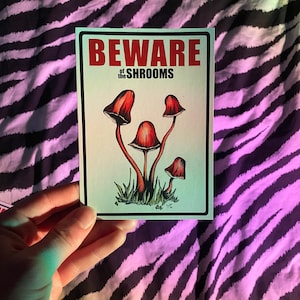 May include: A rectangular sticker featuring a white background with a gradient light blue border. The text BEWARE of the SHROOMS is printed in red and black. Below the text are four red mushrooms with dark stems growing from green grass.