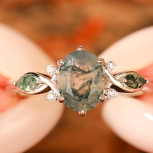 Puede incluir: Un anillo de plata con una piedra preciosa de ágata musgosa de corte ovalado, flanqueada por dos piedras verdes en forma de hoja. El anillo está adornado con pequeñas piedras preciosas redondas y transparentes. El anillo es sostenido por una persona.