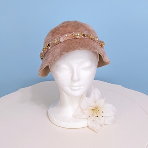 May include: A light pink felt cloche hat with a decorative band of faux pearls and shells.