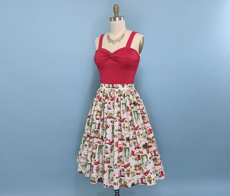 May include: A red, strapless top with a twist detail at the front, paired with a white skirt featuring a vintage-inspired print of kitchen appliances and food. The skirt is full and has a gathered waist.