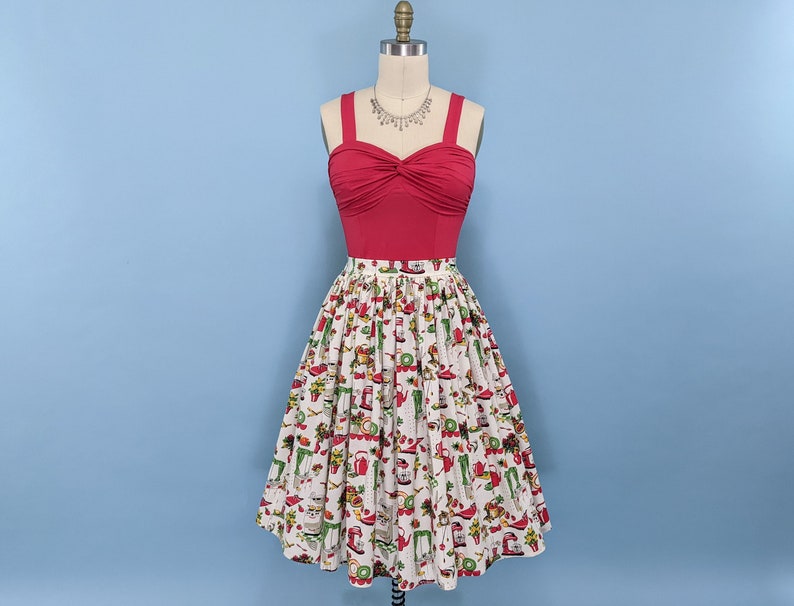 May include: A red sleeveless top with a gathered neckline and a white skirt with a colourful print of kitchen appliances and food. The skirt is full and has a gathered waist.