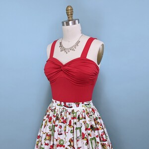 May include: A red sleeveless top with a twist detail at the bust, paired with a white skirt featuring a colourful pattern of kitchen appliances. The top has a silver necklace with a pendant.