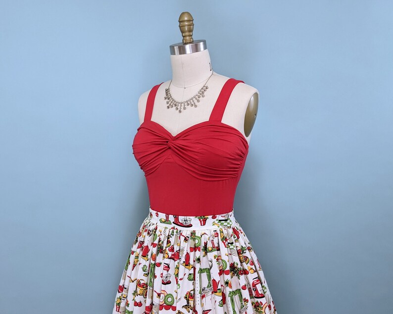 May include: A red sleeveless top with a twist detail at the bust, paired with a white skirt featuring a colourful pattern of kitchen appliances. The top has a silver necklace with a pendant.