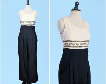May include: A black and white jumpsuit with a gold and black patterned band around the waist. The jumpsuit has a sleeveless top and wide legs.